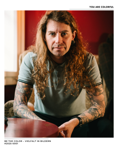 Mann mit langen Haaren und Tattoos sitzt auf einem Stuhl vor roter Wand.